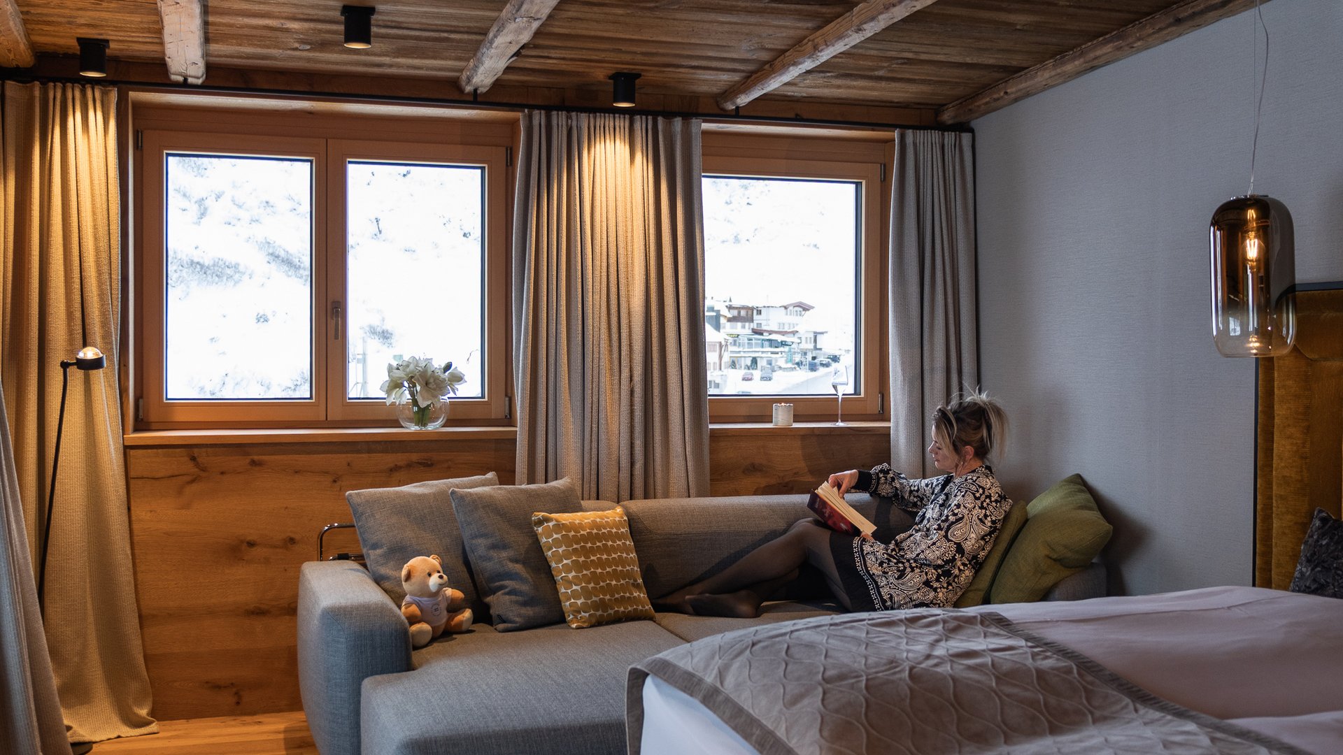 Zum Pressebereich des Hotel Maiensee Frau liest auf grauem Sofa in gemütlichem Holz-Zimmer mit Ausblick auf Schnee