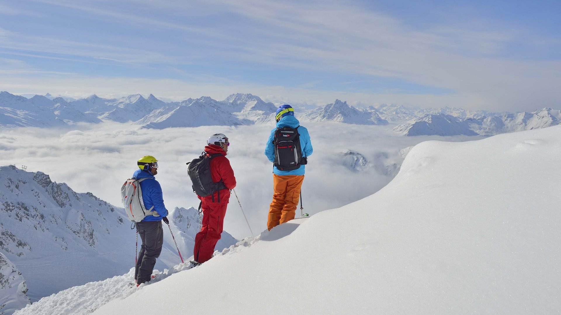 Arlberg: Ihr Hotel an der Piste Drei Skifahrer schauen von verschneitem Berggipfel auf Wolken und Alpen