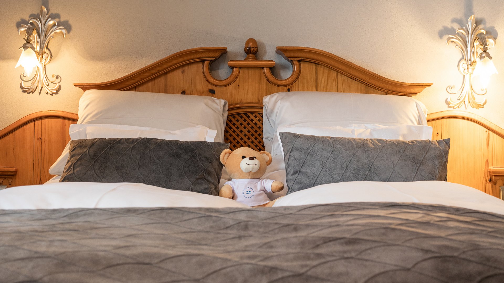 Zum Pressebereich des Hotel Maiensee Gemütliches Bett mit grauen Kissen und einem Teddybär in der Mitte