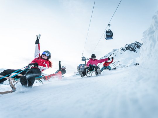 Hotel Maiensee: winter sports at the Arlberg Four people sledding down a snowy mountain with gondolas in the background