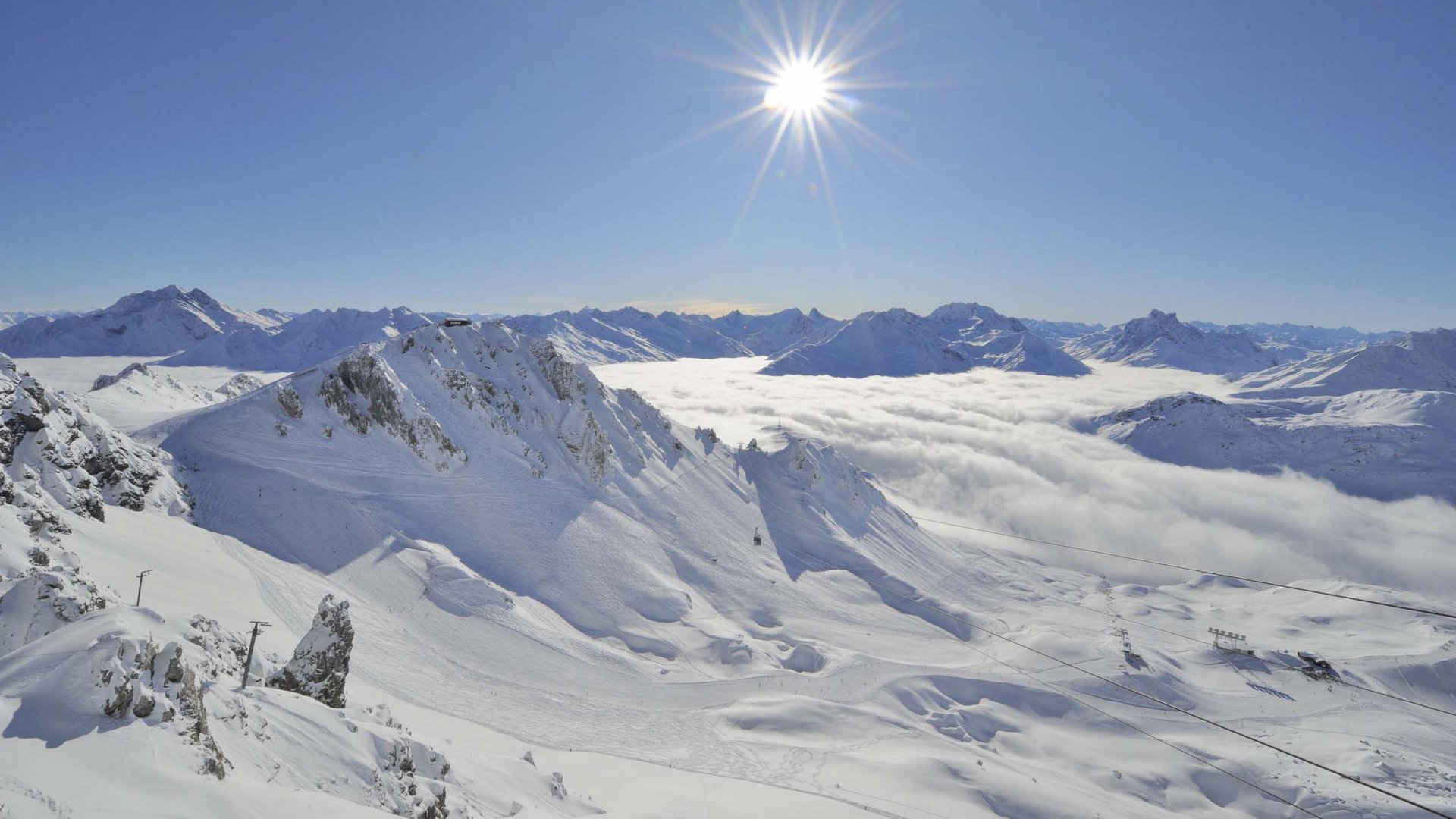 Arlberg: Ihr Hotel an der Piste Sonniger Wintertag mit schneebedeckten Bergen und Wolken im Tal