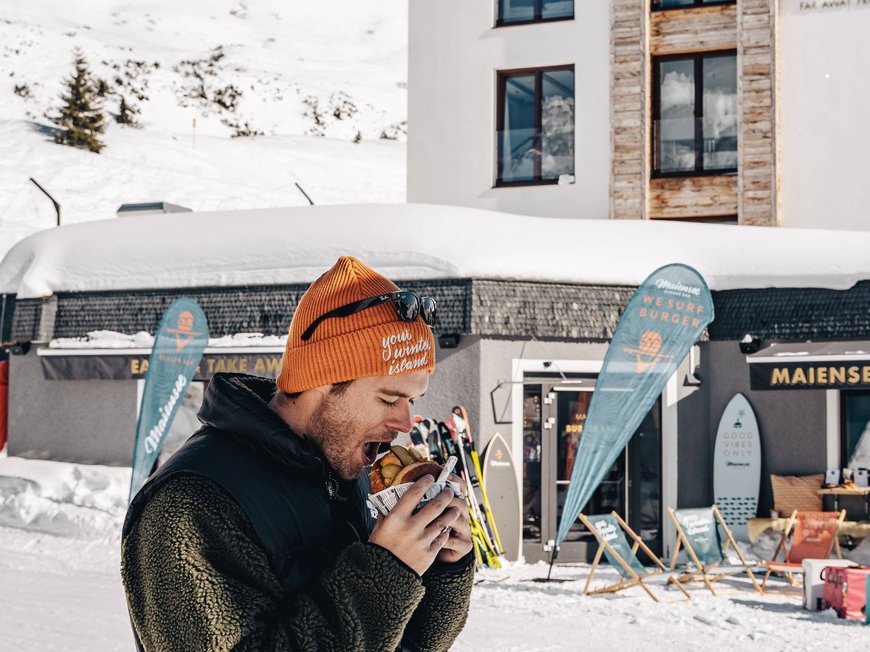 Maiensee, Ihr Hotel in Sankt Christoph: Kulinarik Mann isst Burger vor verschneitem Hotel Maiensee im Winter