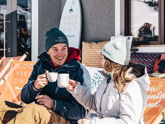 Hotel Maiensee: your hotel in St. Christoph Two people drinking hot beverages outside on snowy terrace