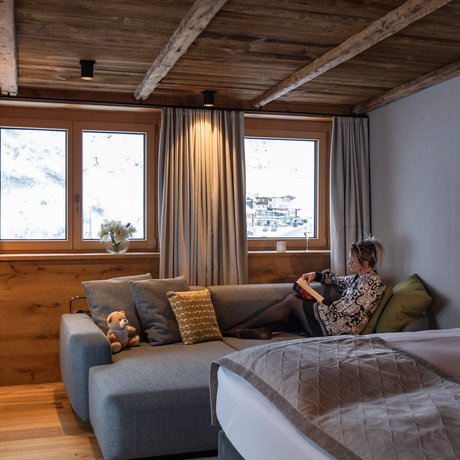 Ihre Unterkunft in St. Christoph am Arlberg: Bilder Frau liest auf Sofa im gemütlichen Zimmer mit Holzbalkendecke und Alpenblick