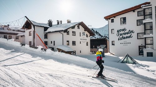 Skiurlaub am Arlberg: Anfrage Skifahrer vor schneebedeckten Hotels in einer sonnigen Berglandschaft