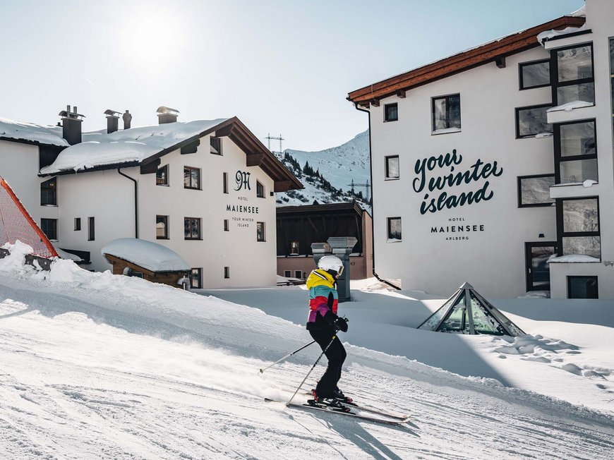 Arlberg: Ihr Hotel an der Piste Skifahrer vor schneebedeckten Hotels in einer sonnigen Berglandschaft