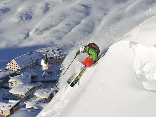 Arlberg: Ihr Hotel an der Piste Skifahrer fährt steilen verschneiten Hang mit Bergdorf im Hintergrund hinab