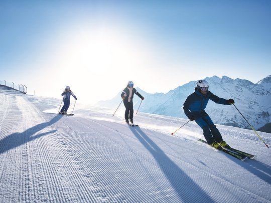 Arlberg: Ihr Hotel an der Piste Drei Skifahrer auf präparierter Piste vor Bergkulisse bei Sonnenschein