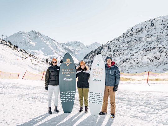 Arlberg: Ihr Hotel an der Piste Drei Personen mit Snowboards vor verschneiten Bergen an einem sonnigen Tag
