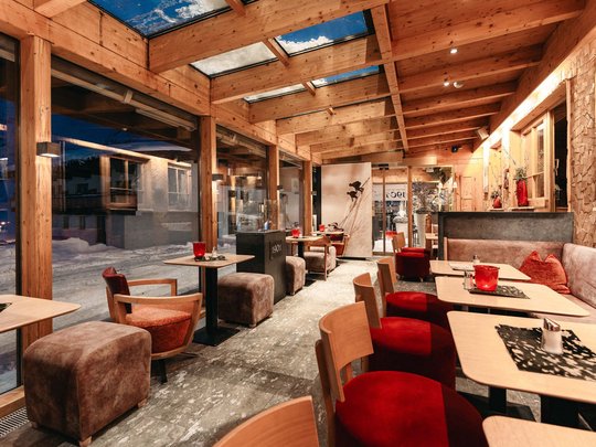 The lounge bar in your ski hotel at the Arlberg Cozy café with wooden beams, large windows, red accents, and seating areas.