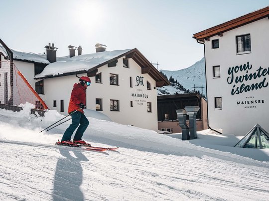 Arlberg: Ihr Hotel an der Piste Skifahrer vor verschneitem Hotel Maiensee in den Bergen am sonnigen Wintertag