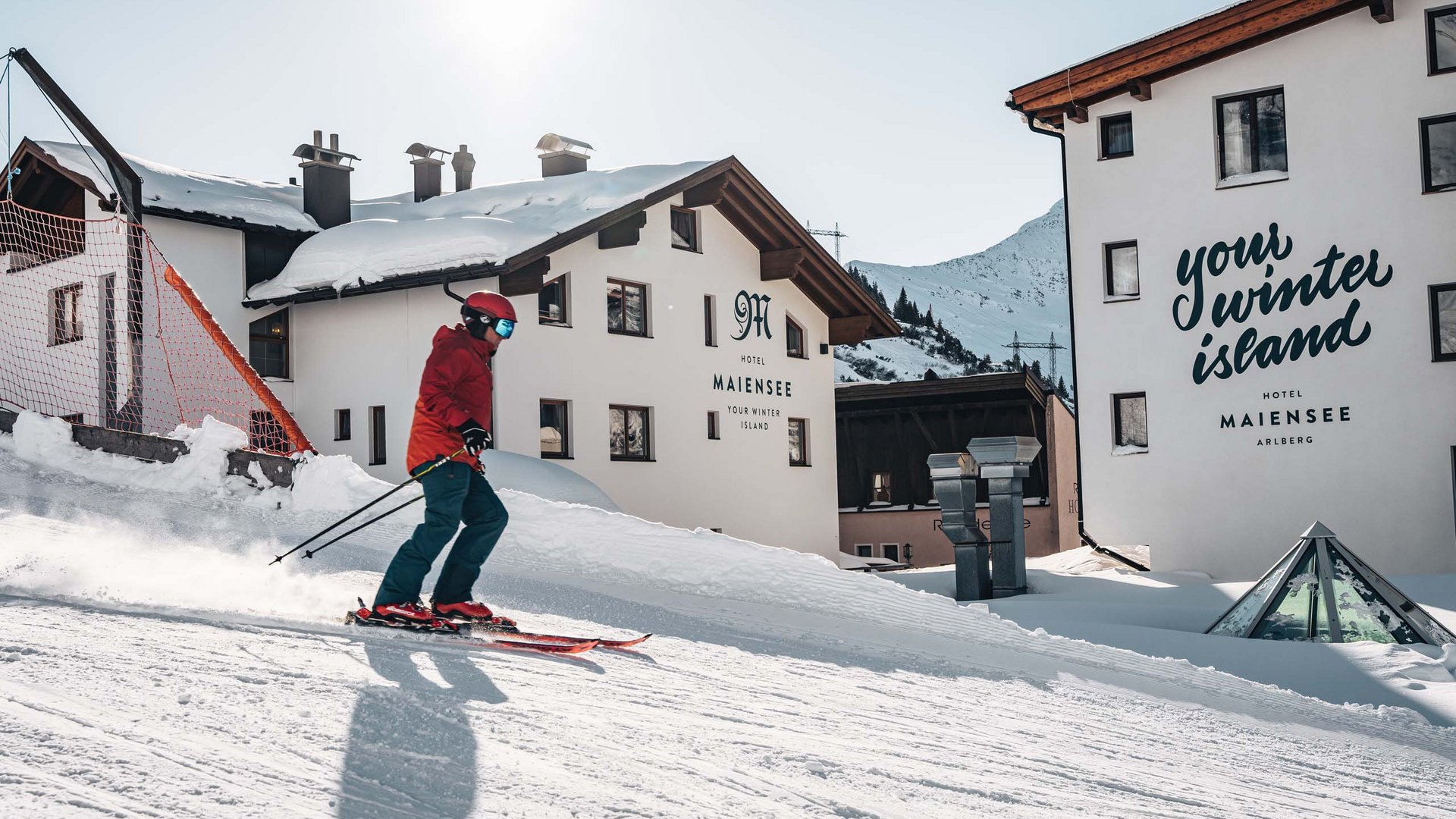 Arlberg: Ihr Hotel an der Piste Skifahrer vor verschneitem Hotel Maiensee in den Bergen am sonnigen Wintertag