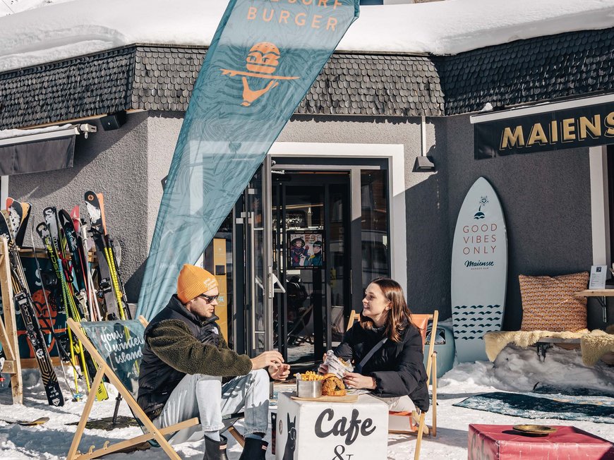 Premium burgers at the ski hotel near St. Anton am Arlberg Two people sitting in the snow outside a cafe and bar, surrounded by skis.